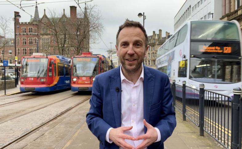 Cllr Ben Miskell stands in Sheffield city centre with two trams parked up behind him on his right hand side and a double decker bus on his left hand side