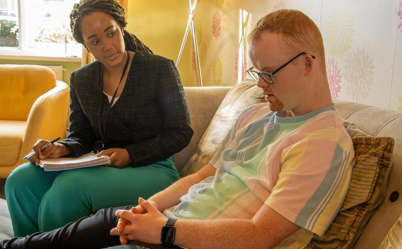 A lady sits with a young man. She is holding a notebook and a pen. He wears glasses and looks down at his hands