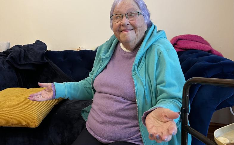 Pat, smiling with her arms raised slightly at her sides sitting on her sofa