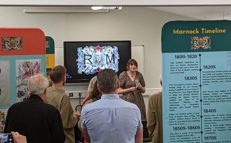 People with their backs to the camera facing a presenter who is talking about the exhibition to her. Exhibition boards in the foreground.