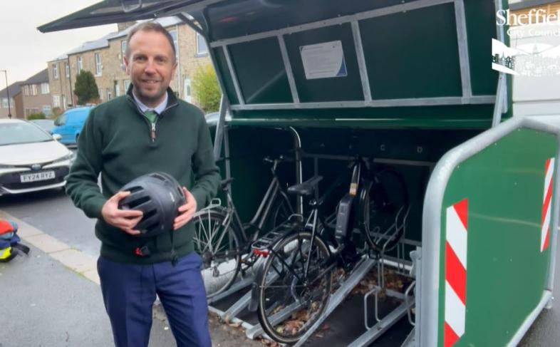Councillor Ben Miskell stands in front of of an open cycle hangar on a busy street, with cars in the background. There are bikes in the hangar, and information is fixed to the inside of the hangar lid.