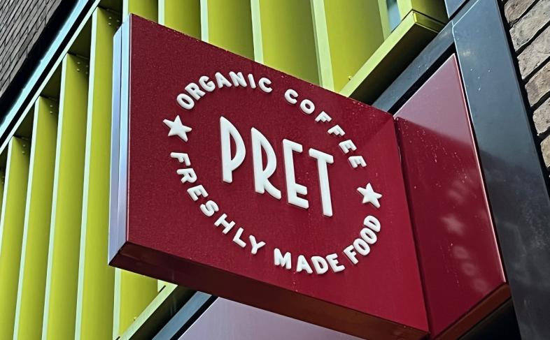 A Pret sign hanging from the side of the new store in Sheffield city centre, the image shows the sign with a yellow, metal, design of the building in the background.