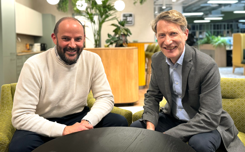 Alexis Krachai, left, and Matthew Nimmo, right, sit on green chairs in a bright office behind a round table, with plants and bright hanging lights in the background