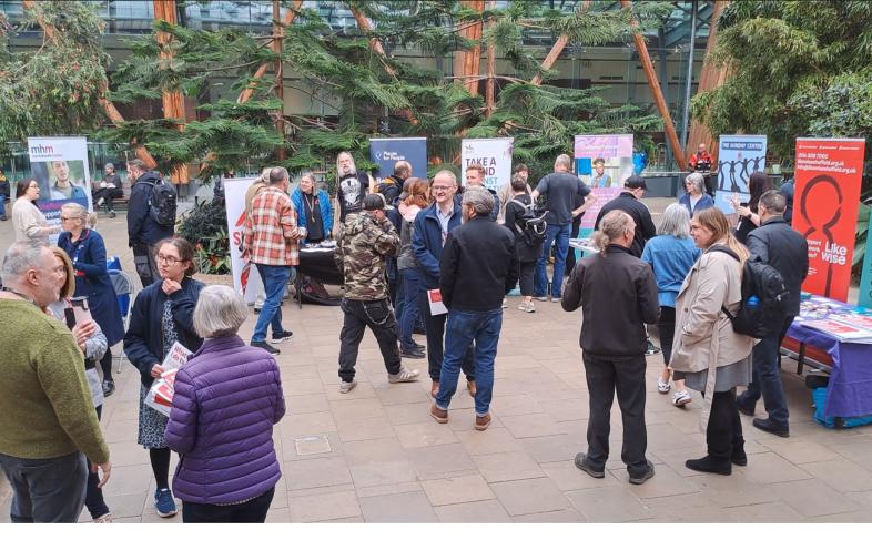 Attendees at the Help Us Help launch event in The Winter Garden chatting and networking in front of stalls displaying partner information as the public walk past