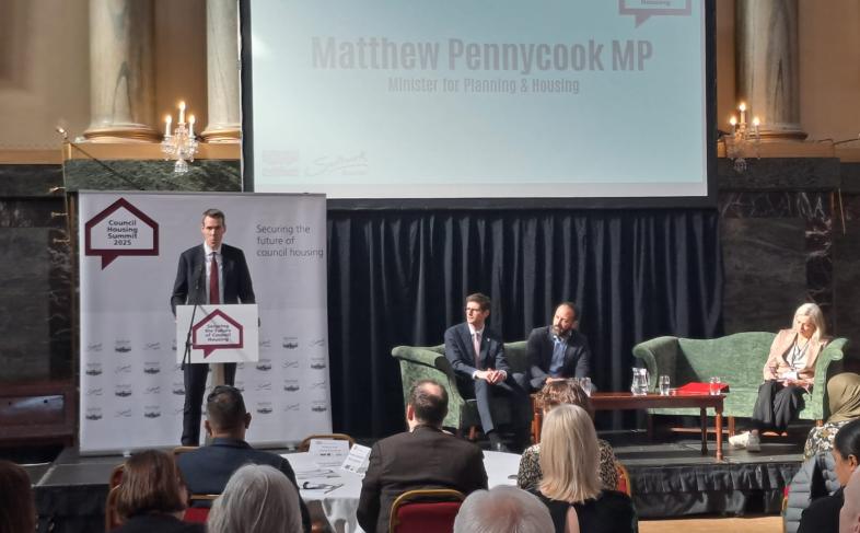 Matthew Pennycook MP addresses a room full of delegates at Sheffield's Cutler's Hall on a stage, while Cllr Tom Hunt, Cllr Kieron Williams and Kate Josephs watch on, sat on safas in front of a screen showing the speaker's name