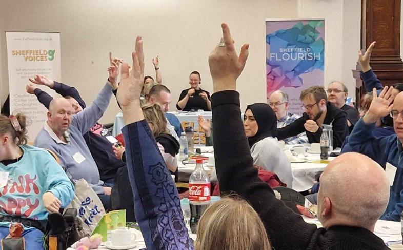 Participants at Big Voices event with some hands raised at some of the tables