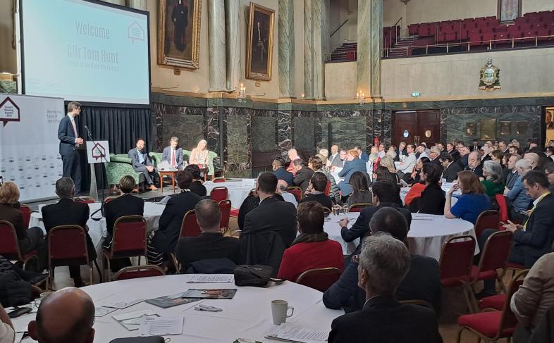 Sheffield City Council Leader, Cllr Tom Hunt, stands at a podium on stage at Cutler's Hall in Sheffield delivering a speech to a packed audience. Behind him sat on a sofa on the stage are Southwark Council Leader Cllr Kieron Williams, Housing Minister Matthew Pennycook and Sheffield City Council Chief Executive Kate Josephs