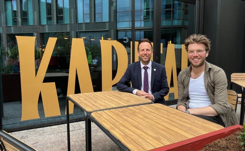 Cllr Ben Miskell sits at a table with Ben Stubbs from Kapital Bar, behind them is Kapital written in big green letters on the window of the bar