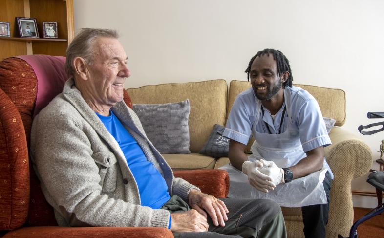 A man sitting in an armchair and a carer sits on the sofa. Both are smiling.
