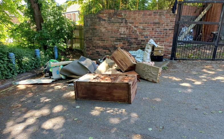 Fly-tipped waste on the corner of a round with a brick wall and trees in the background