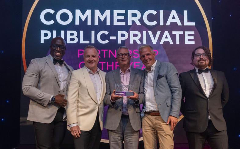 Five people stand on a stage in front of a projector screen with the words Commercial Public-Private written on it, the person in the middle of the group is holding an award