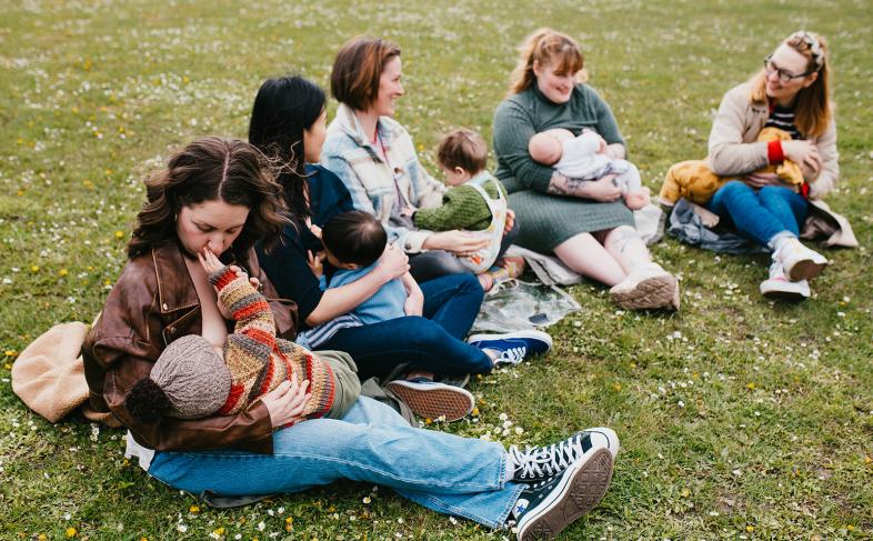 Parents breastfeeding and socialising while sat on a grassy field. 