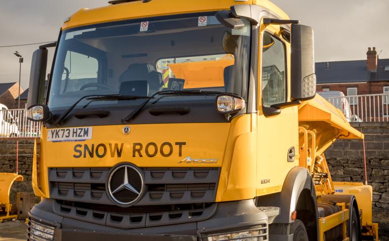 Image is of a yellow grit lorry with the name 'Snow Root' written on the front of it.