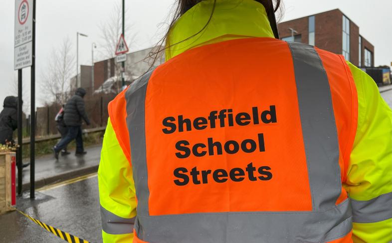 A person stands on a street wearing a high visibility jacket with the words School Street written on the back