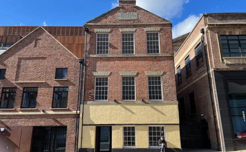Picture shows the front of the old Sunday School building on Cambridge Street, the building is three floors tall with a total of eight windows and a door, a man sits on the edge of the building on the ground floor