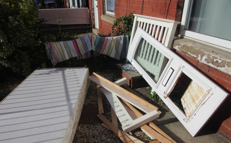 A number of damaged household items such as a bed and mattress and a broken door are strewn messily around a front garden of a house on a bright, sunny day
