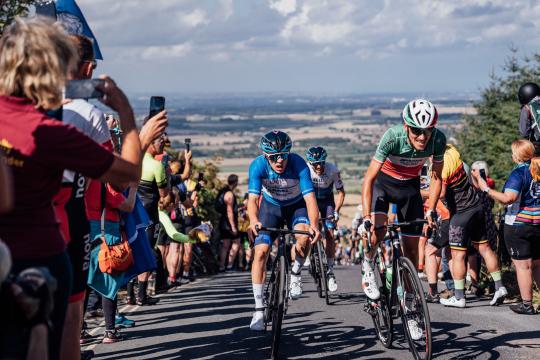 Action from the Tour of Britain, cyclists climbing a hill with spectators surrounding them, with hills and countryside in the distance 