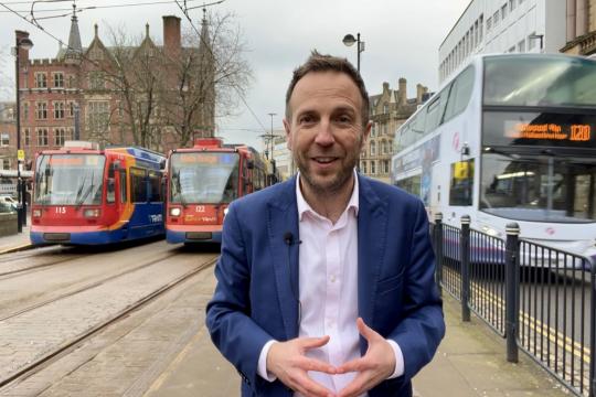 Cllr Ben Miskell stands in Sheffield city centre with two trams parked up behind him on his right hand side and a double decker bus on his left hand side