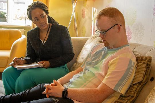 A lady sits with a young man. She is holding a notebook and a pen. He wears glasses and looks down at his hands