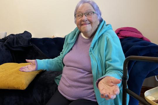 Pat, smiling with her arms raised slightly at her sides sitting on her sofa