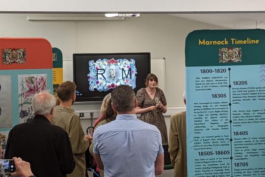 People with their backs to the camera facing a presenter who is talking about the exhibition to her. Exhibition boards in the foreground.