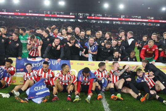 Members of the Sheffield United squad are stood and knelt down on the Bramall Lane pitch while celebrating promotion to the Premier League