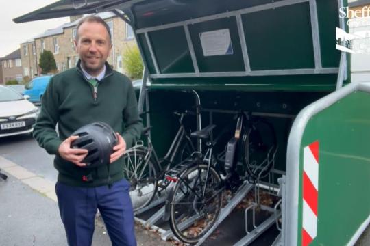 Councillor Ben Miskell stands in front of of an open cycle hangar on a busy street, with cars in the background. There are bikes in the hangar, and information is fixed to the inside of the hangar lid.