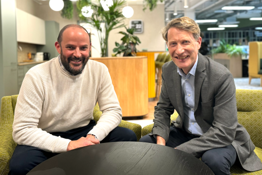 Alexis Krachai, left, and Matthew Nimmo, right, sit on green chairs in a bright office behind a round table, with plants and bright hanging lights in the background