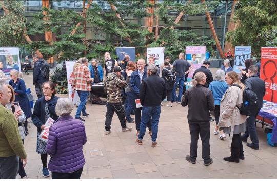 Attendees at the Help Us Help launch event in The Winter Garden chatting and networking in front of stalls displaying partner information as the public walk past