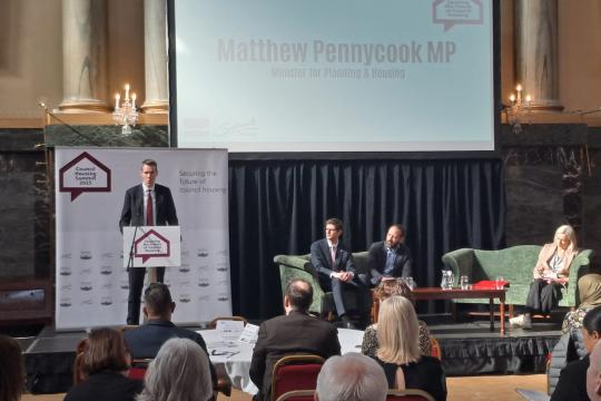 Matthew Pennycook MP addresses a room full of delegates at Sheffield's Cutler's Hall on a stage, while Cllr Tom Hunt, Cllr Kieron Williams and Kate Josephs watch on, sat on safas in front of a screen showing the speaker's name