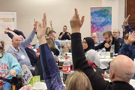 Participants at Big Voices event with some hands raised at some of the tables