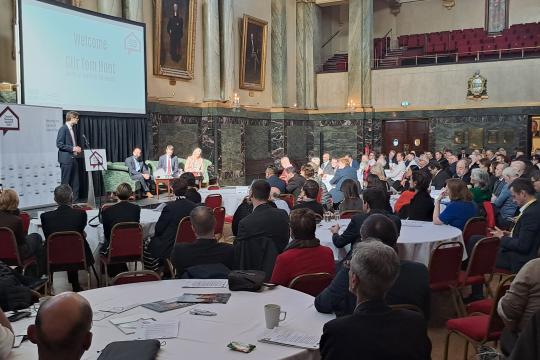 Sheffield City Council Leader, Cllr Tom Hunt, stands at a podium on stage at Cutler's Hall in Sheffield delivering a speech to a packed audience. Behind him sat on a sofa on the stage are Southwark Council Leader Cllr Kieron Williams, Housing Minister Matthew Pennycook and Sheffield City Council Chief Executive Kate Josephs