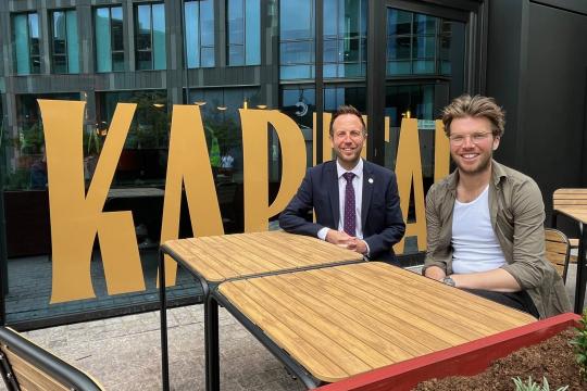 Cllr Ben Miskell sits at a table with Ben Stubbs from Kapital Bar, behind them is Kapital written in big green letters on the window of the bar