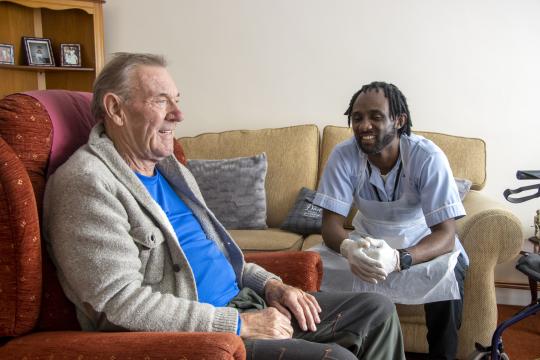 A man sitting in an armchair and a carer sits on the sofa. Both are smiling.