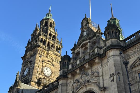 A view of the Sheffield Town Hall clock tower taken from street level looking up. The Union flag can be seen flying from the pole next to the clock tower.