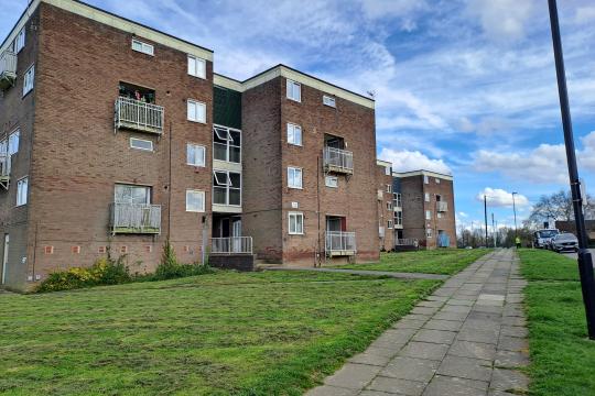 Brown, rendered housing blocks surrounded by grass and a pathway running past them on a sunny day with clouds in the blue sky
