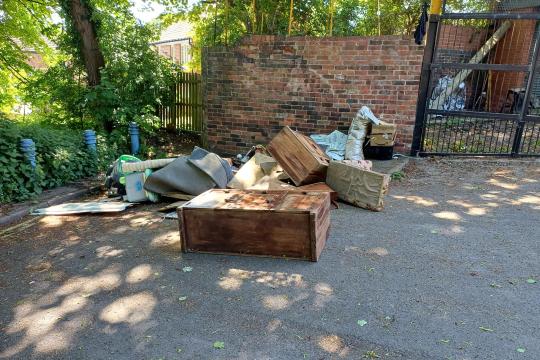 Fly-tipped waste on the corner of a round with a brick wall and trees in the background