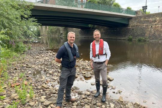 Two people stand on the banks of river with vegetation to the left hand side of them and a bridge going over the river in the background