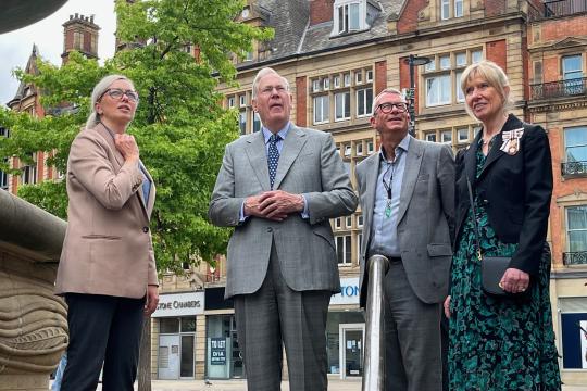 Four people are pictured standing all looking to their left hand sides at something high up, behind them is a tree and some buildings