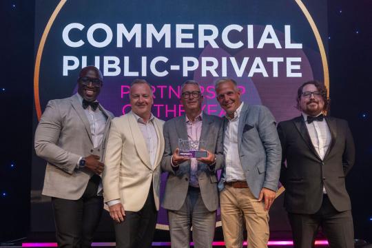 Five people stand on a stage in front of a projector screen with the words Commercial Public-Private written on it, the person in the middle of the group is holding an award