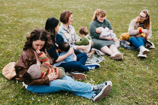 Parents breastfeeding and socialising while sat on a grassy field. 