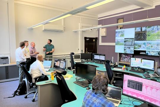 People standing up and sitting down at desk in a large white room with a bank of TV screens on the right hand side wall