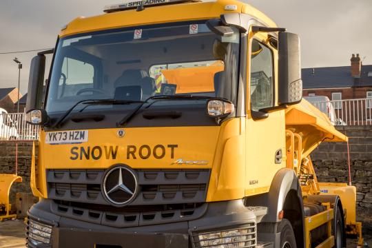 Image is of a yellow grit lorry with the name 'Snow Root' written on the front of it.