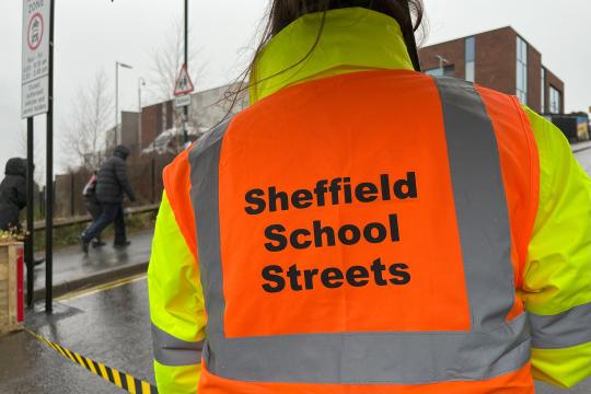 A person stands on a street wearing a high visibility jacket with the words School Street written on the back