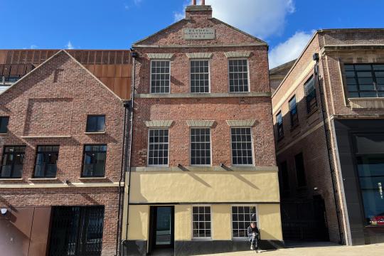 Picture shows the front of the old Sunday School building on Cambridge Street, the building is three floors tall with a total of eight windows and a door, a man sits on the edge of the building on the ground floor