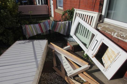 A number of damaged household items such as a bed and mattress and a broken door are strewn messily around a front garden of a house on a bright, sunny day