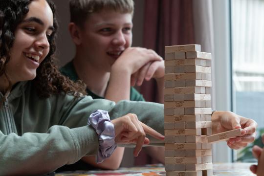 Two children are laughing while sat at a glass table playing a game of Jenga together. There are dark curtains behind them in the background