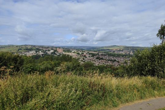 A view from the highest path at Bole Hill Recreation Ground. Housing can be seen in the distance, with plenty of green vegetation in the foreground. There is a mixture of blue skies and clouds above.