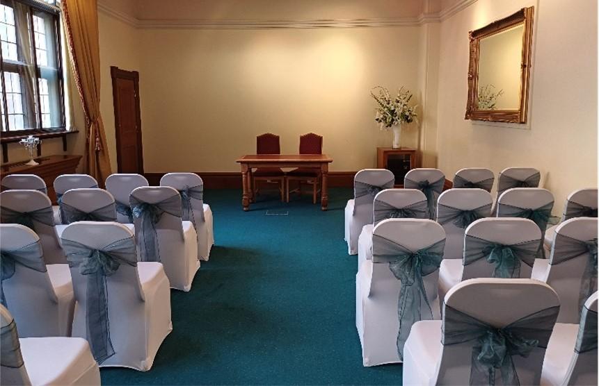 Two rows of seats dressed in white with pale blue bows tied at the back with a central aisle face a table with two chairs facing the rows. A small table to the right of the top table holds a vase with a floral arrangement. A gilded mirror is mounted on the right hand wall. A window on the left hand wall with gold curtains tied back.